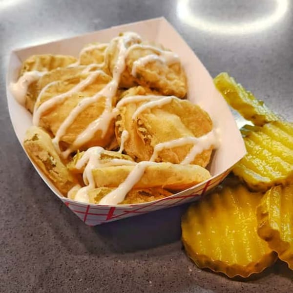Fried pickles drizzled with white sauce in a paper tray, with pickle slices on the side.
