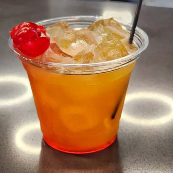 A vibrant orange drink with ice and two red cherries in a clear plastic cup.