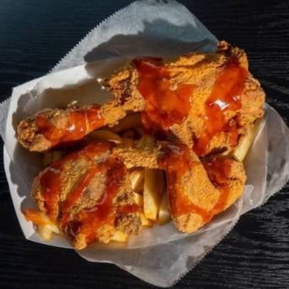 Crispy fried chicken pieces with hot sauce and french fries in a white paper tray.