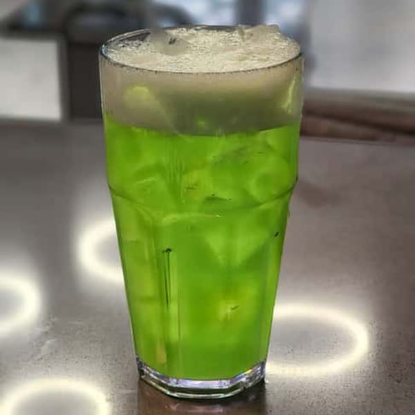 A tall glass of bright green soda with ice and a foamy top sits on a reflective surface.