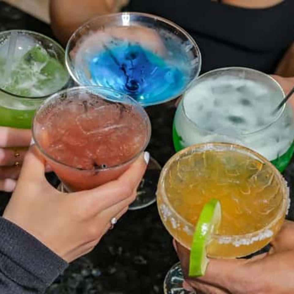 Five hands holding colorful cocktails, including blue, green, red, and yellow drinks, some with lime.