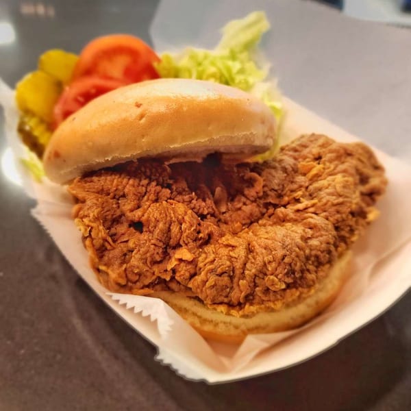 Crispy fried chicken sandwich with lettuce, tomato, and pickles on a bun in a paper tray.